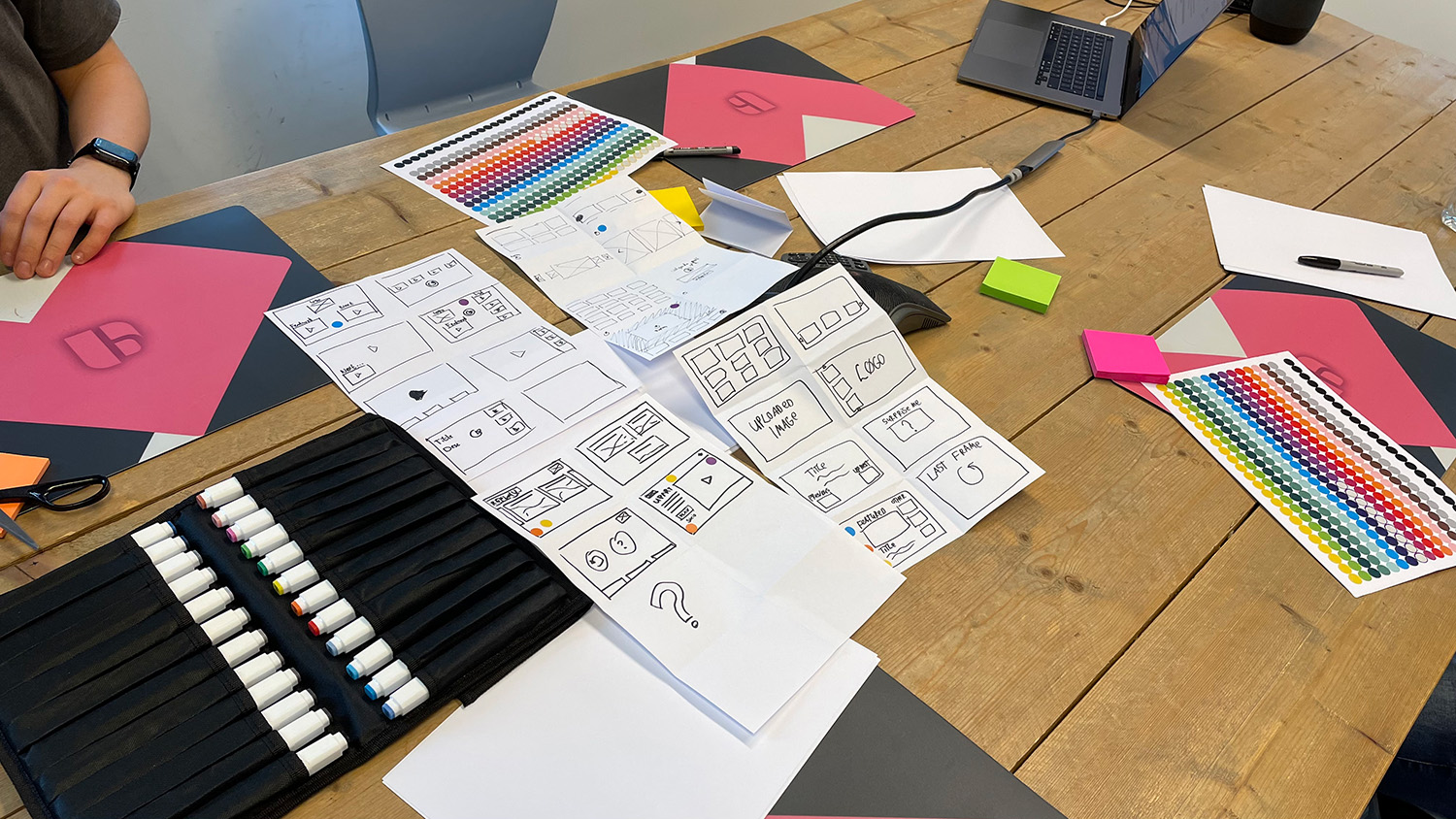 Folded sheets of paper with sketches of player layouts on a table, next to markers and colour stickers.
