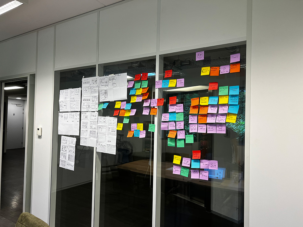 A glass wall with hand-drawn interface sketches on paper next to groups of coloured sticky notes.