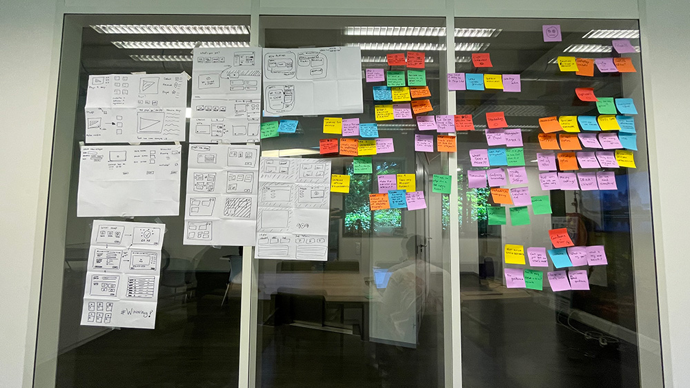 A glass wall with paper wireframe sketches and many coloured sticky notes.